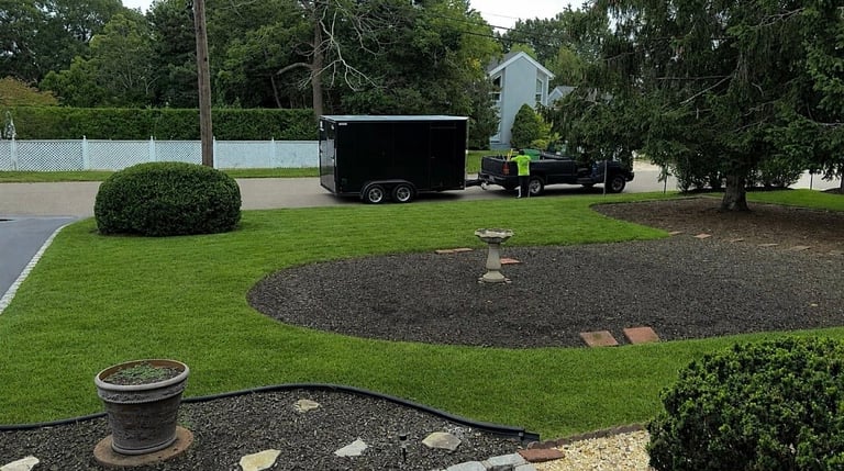 Residential driveway with black enclosed trailer parked behind a white fence, surrounded by manicured lawn and landscaped garden beds