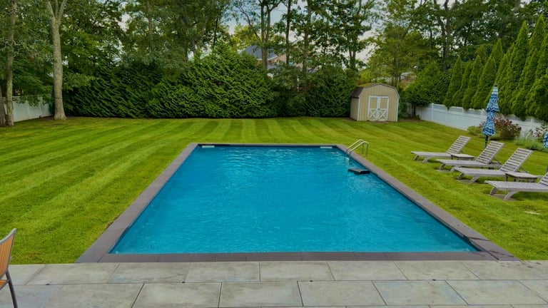 A rectangular turquoise swimming pool surrounded by manicured green lawn, with lounge chairs and garden structures visible in the backyard