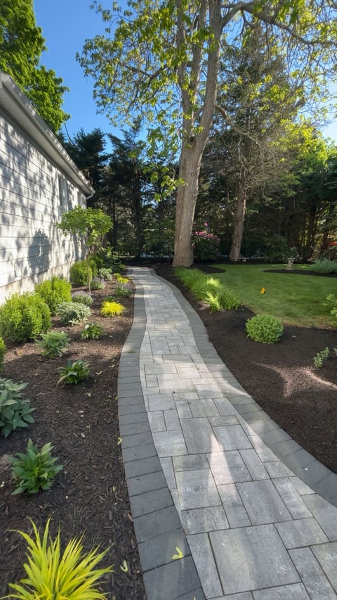 Stone paver pathway leading through a landscaped garden with mature trees, manicured lawn, and green plantings alongside a shingled house