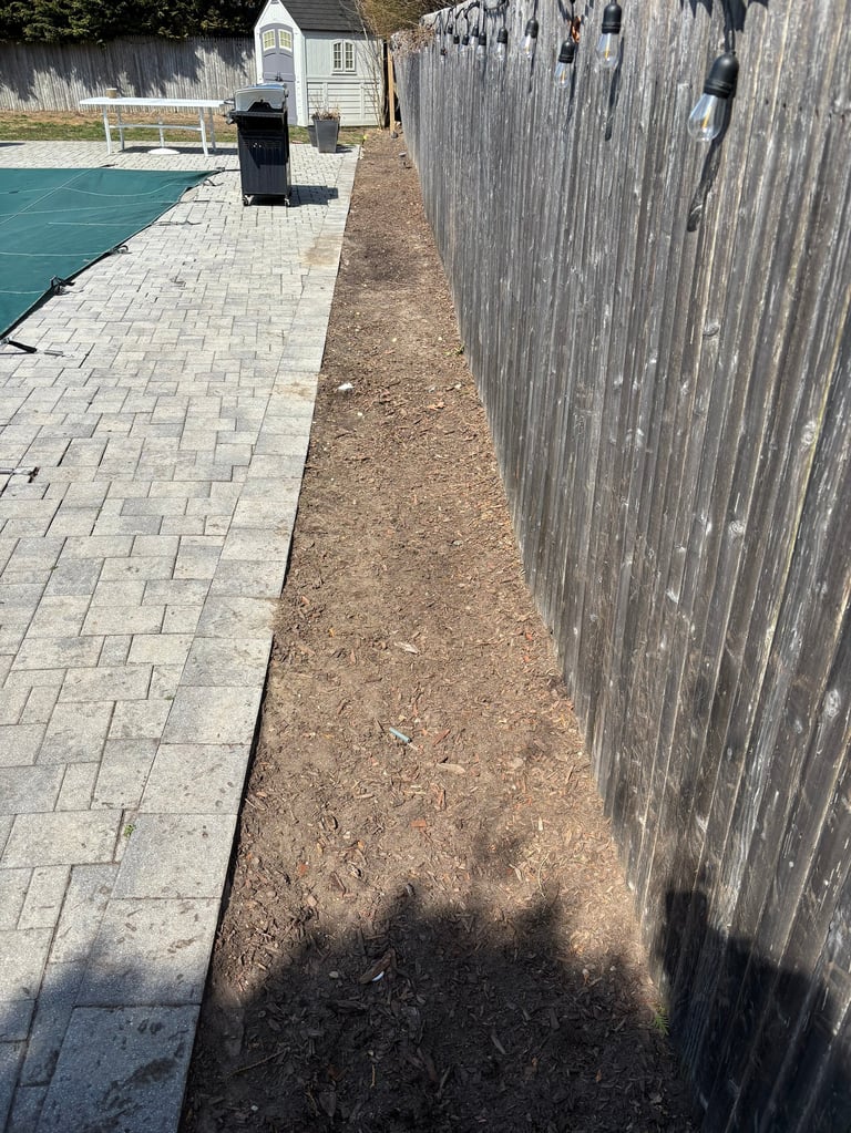 Narrow walkway between weathered wooden fence and paver patio beside a swimming pool with pool house