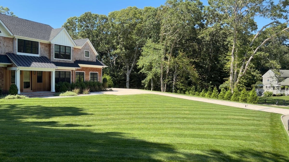 Modern two-story brick and stone house with large manicured lawn and mature trees in background on sunny day