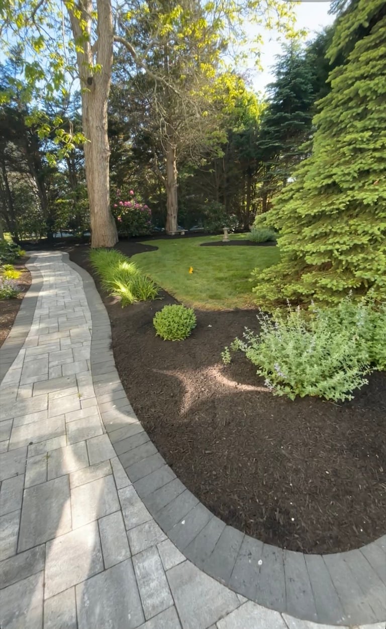 Curved stone patio pathway winds through a manicured garden with tall trees, ivy-covered hedge, and lush green lawn