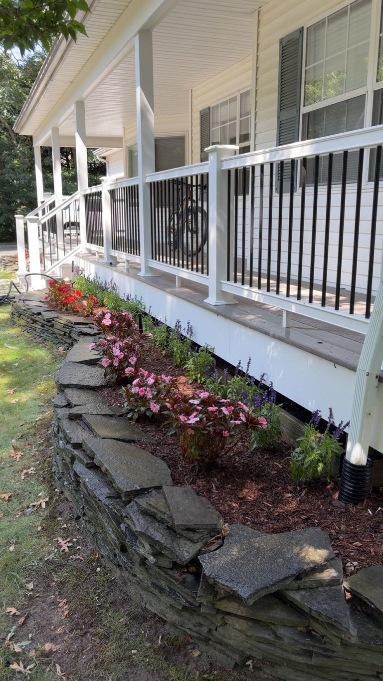 Front porch of white house with black and white railings, landscaped flower bed with pink flowers and stone border