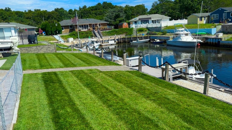 Waterfront property with manicured lawn, docked boats, residential homes, and tree-lined background on sunny day