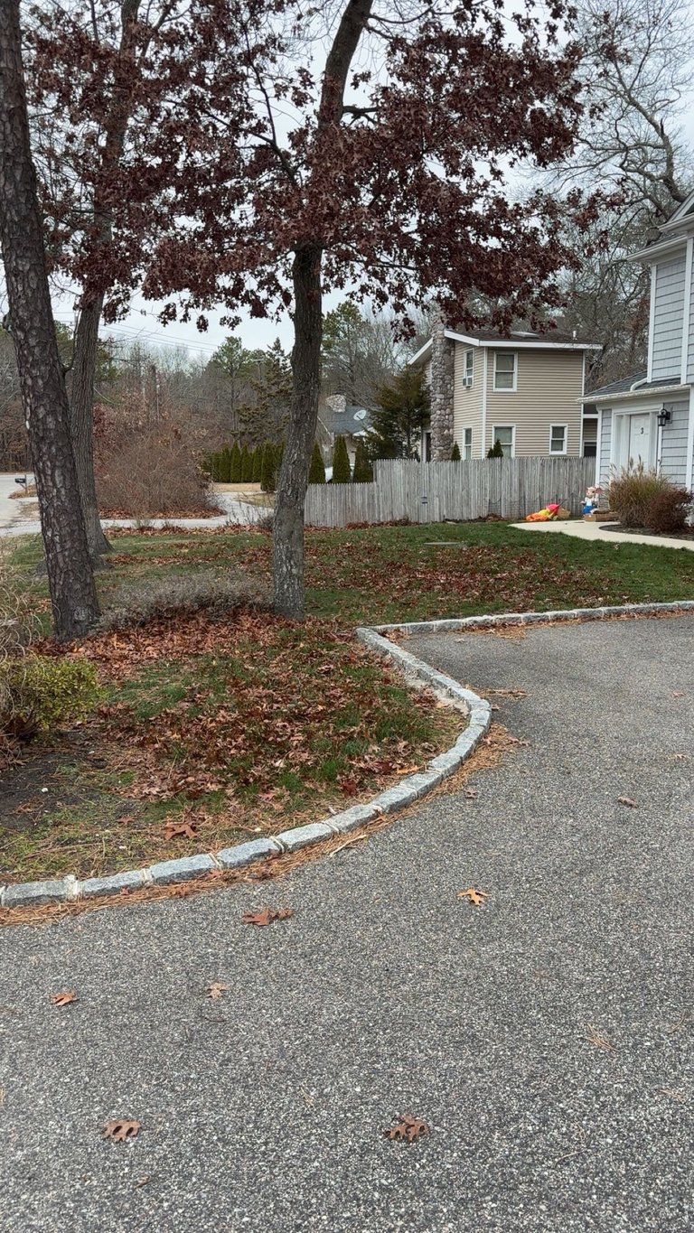 Suburban home with curved driveway, large bare trees, brown leaf-covered lawn, and white wooden fence in fall season