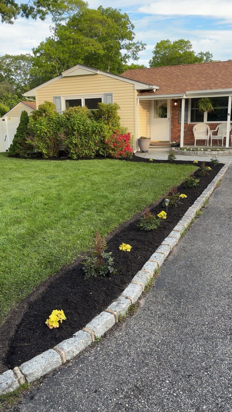 Suburban single-story home with cream and brick exterior, manicured lawn, black mulch garden bed with yellow flowers lining the driveway, mature trees, and covered porch