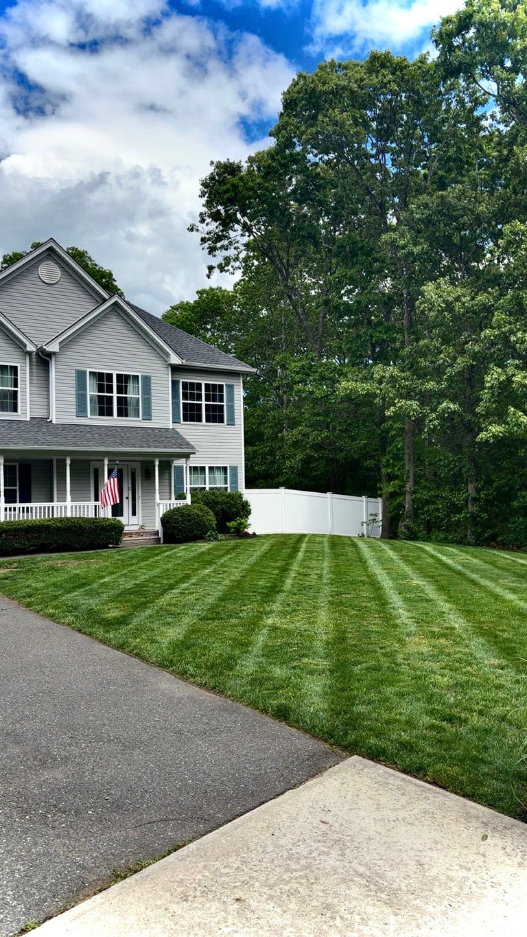Two-story white farmhouse with manicured lawn, striped grass pattern, and mature trees against blue sky with clouds