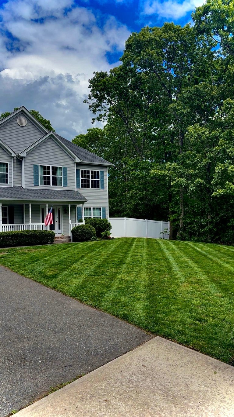 Two-story white house with manicured lawn and striped grass pattern, American flag on porch, surrounded by tall green trees under blue sky