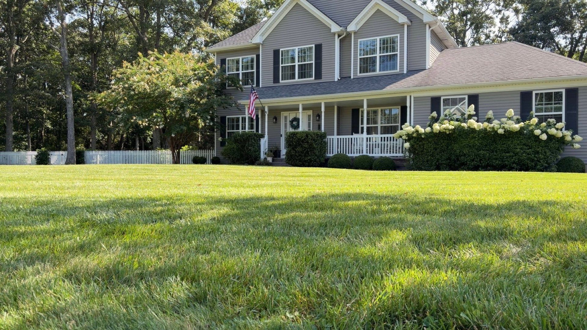 Gray two-story house with white trim and front porch, surrounded by well-maintained lawn and mature trees