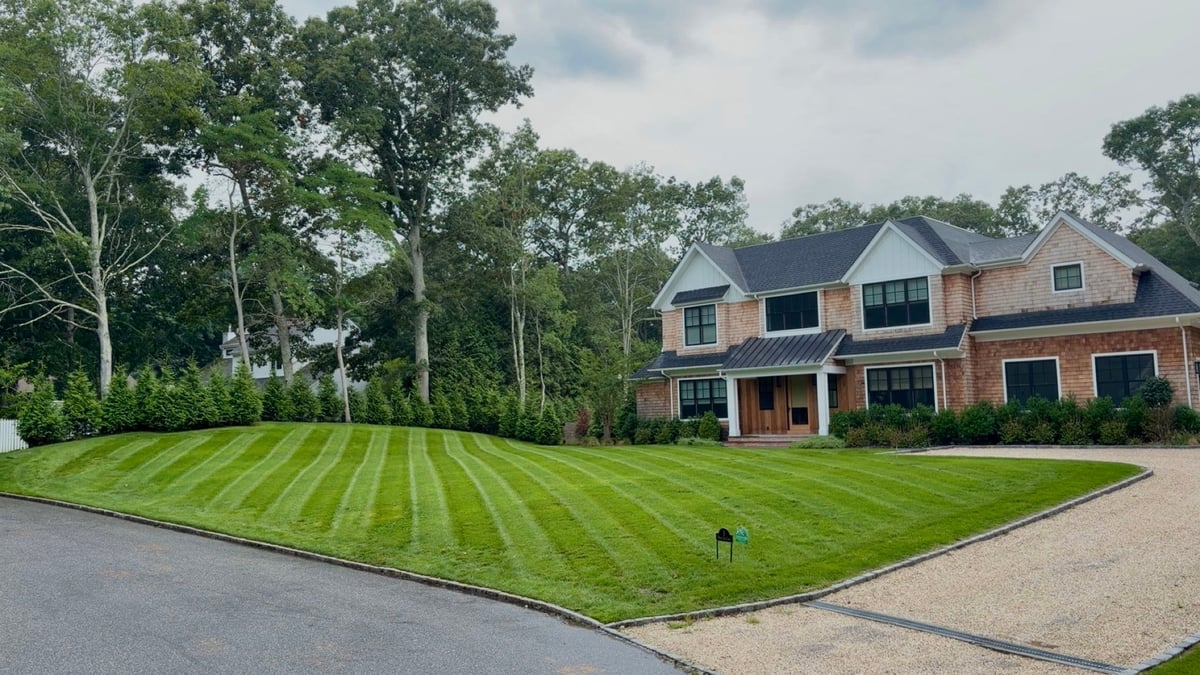 Large brick two-story house with manicured striped lawn and driveway, surrounded by tall trees on an overcast day