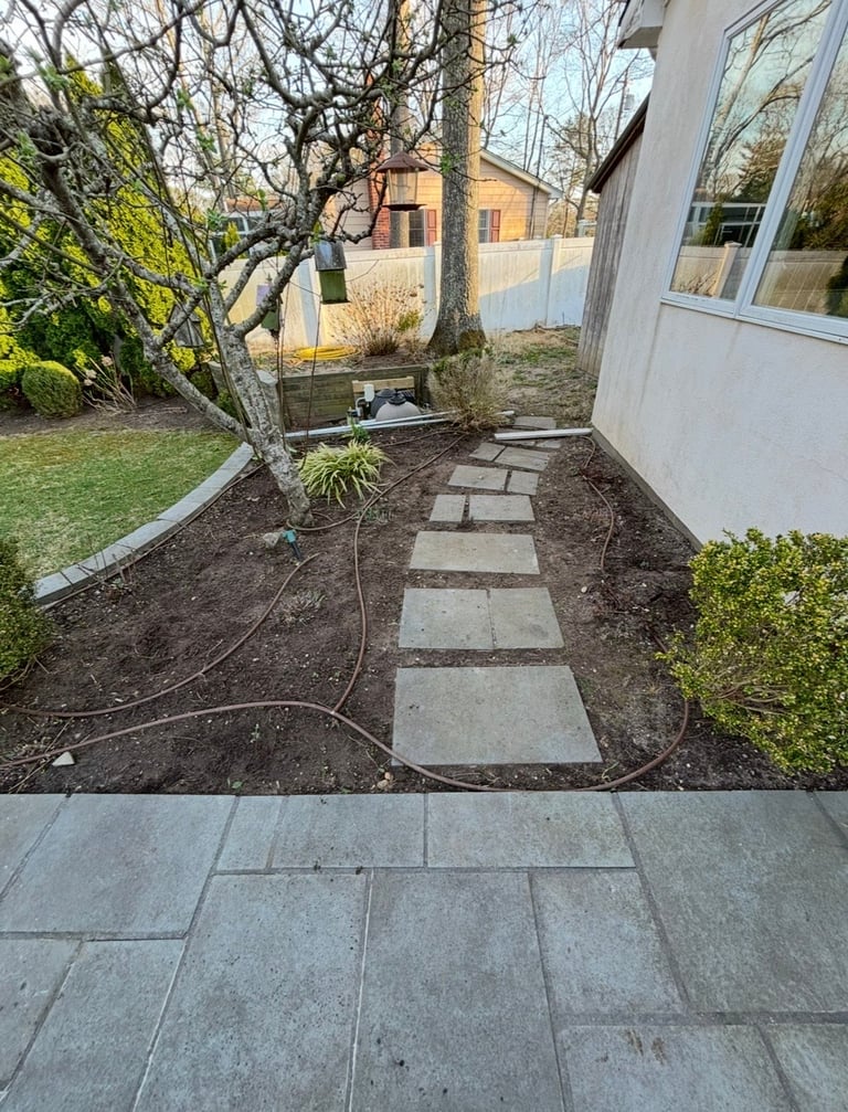 Narrow backyard garden pathway with stepping stones leading between patio and white building, surrounded by dormant trees and shrubs